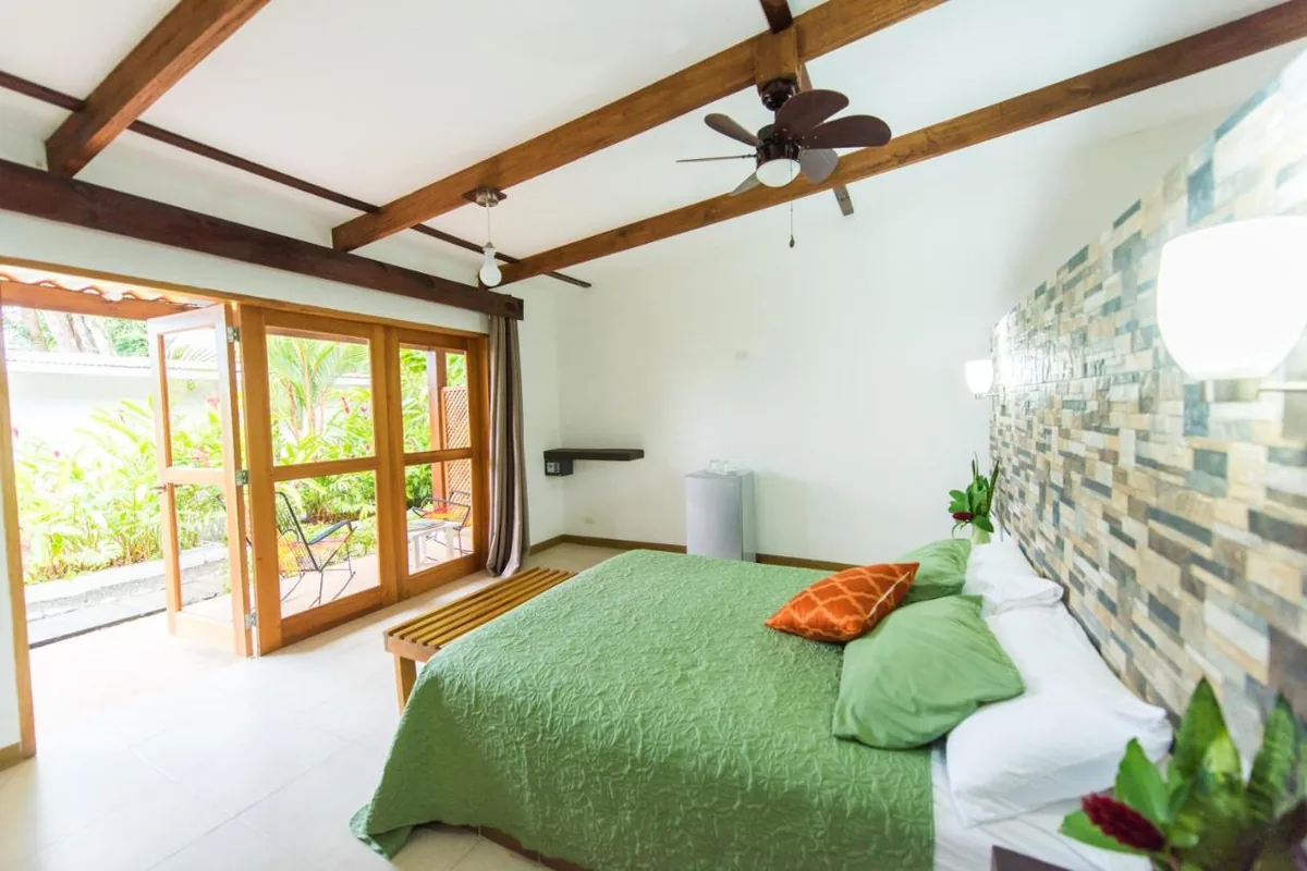 Habitación con pared de piedra, cama con cobija verde y puertas de madera y vidrio abiertas al jardín.